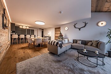 Cozy, modern open-plan living area with a gray sectional sofa, dining table, kitchen bar, wood accents, and a festive Christmas tree.