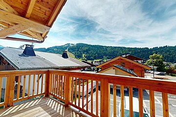 View from a sunny wooden balcony overlooking a village with chalets, surrounded by forested mountains under a bright blue sky.