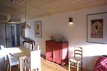 A rustic dining room with a light wood table, white chairs, red cabinet, and framed art under a wooden ceiling, warmly lit by sunlight.