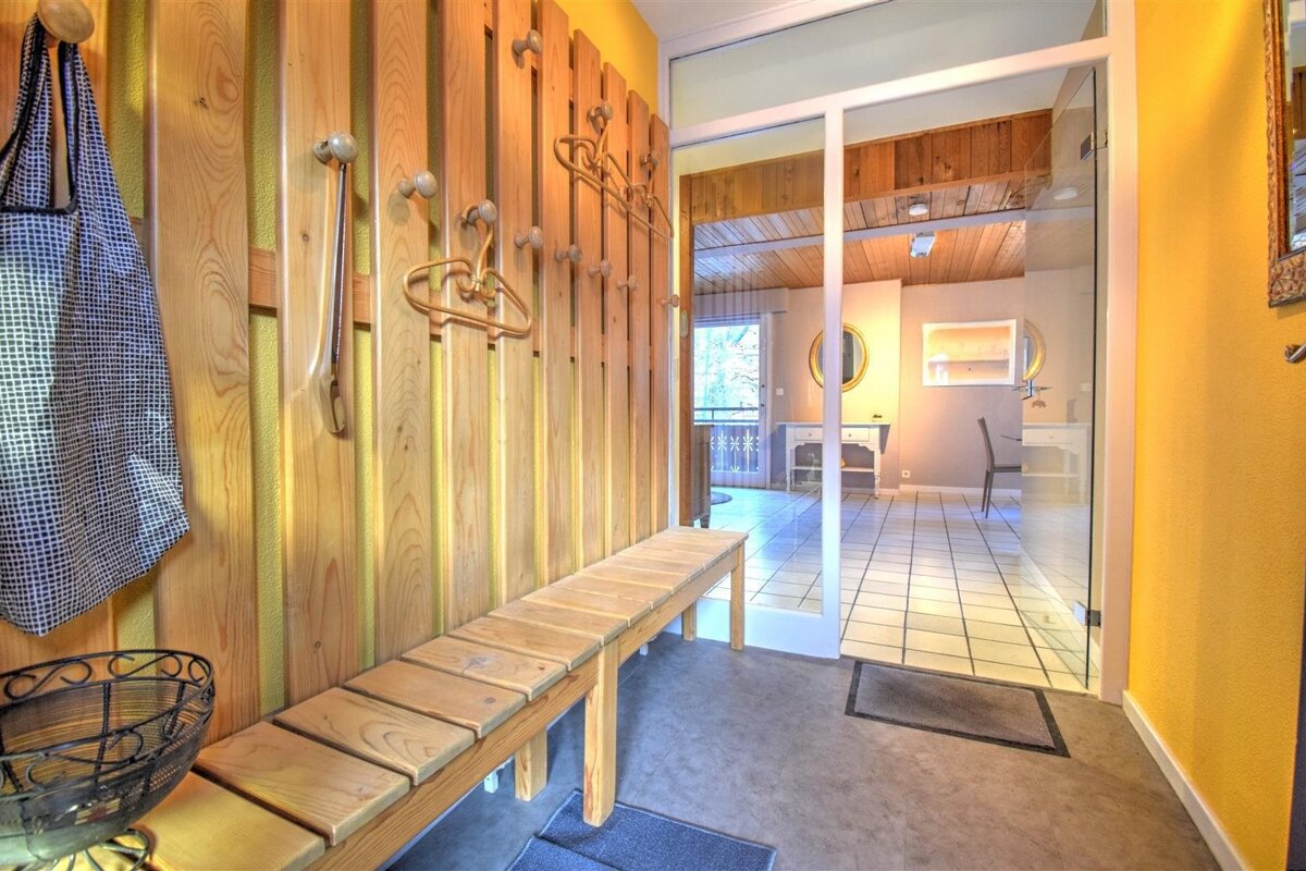Bright entryway with a wooden coat rack, bench, and yellow walls. A glass door opens to a tiled room with mirrors and a balcony view.
