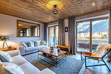 Modern chalet living room with warm wood ceiling, comfy seating, unique lamp, and large windows overlooking snowy mountains.