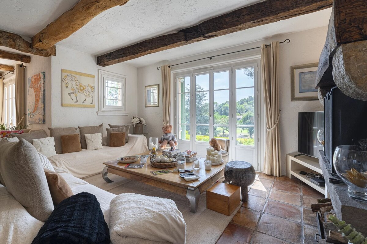 A cozy, rustic living room featuring exposed wooden ceiling beams, cream sofas, and French doors that open to a scenic green view.