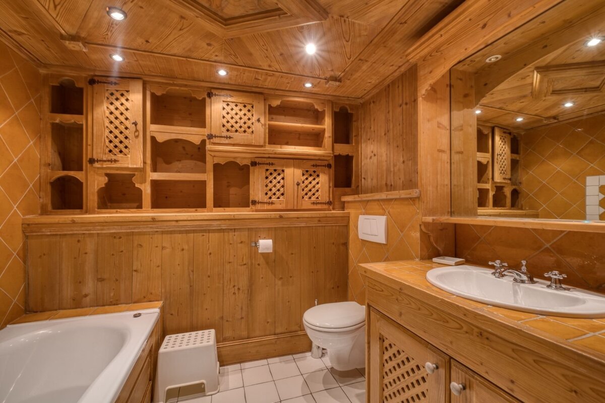 A warm, rustic bathroom features extensive light wood paneling on walls and ceiling, orange diamond tiles, a bathtub, toilet, and a sink with a wooden vanity.