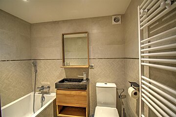 Modern beige-tiled bathroom featuring a bathtub, wooden vanity with a black stone sink, white toilet, and a heated towel rail.