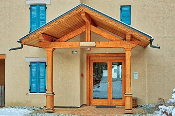 A beige building features a wooden porch over glass doors, flanked by windows with bright blue shutters. Light snow covers the ground and porch roof.