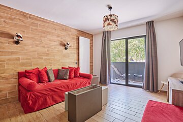 A modern living room featuring a vibrant red sofa, wood-paneled wall, cow print lighting fixtures, light wooden floor, and a balcony with greenery.