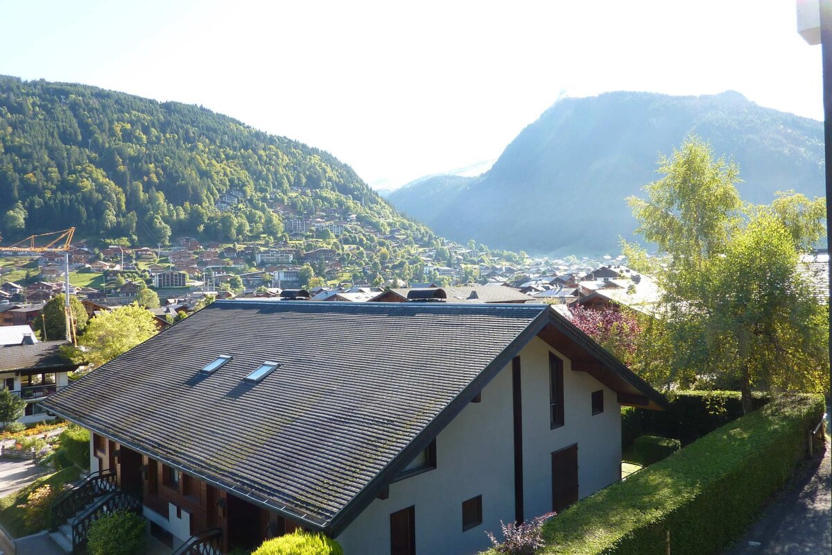 A house roof in the foreground, overlooking a sunny mountain village nestled in a lush green valley with forested hills and hazy peaks.