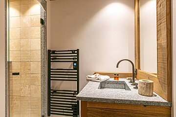 Modern bathroom with a stone-tiled walk-in shower, dark heated towel rack, and a unique stone sink with a wooden mirror frame.