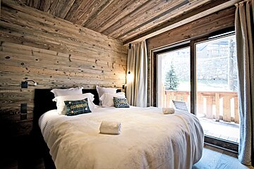Chalet-style bedroom with a large white bed, wood-paneled walls/ceiling, and a glass door opening to a sunny outdoor balcony.