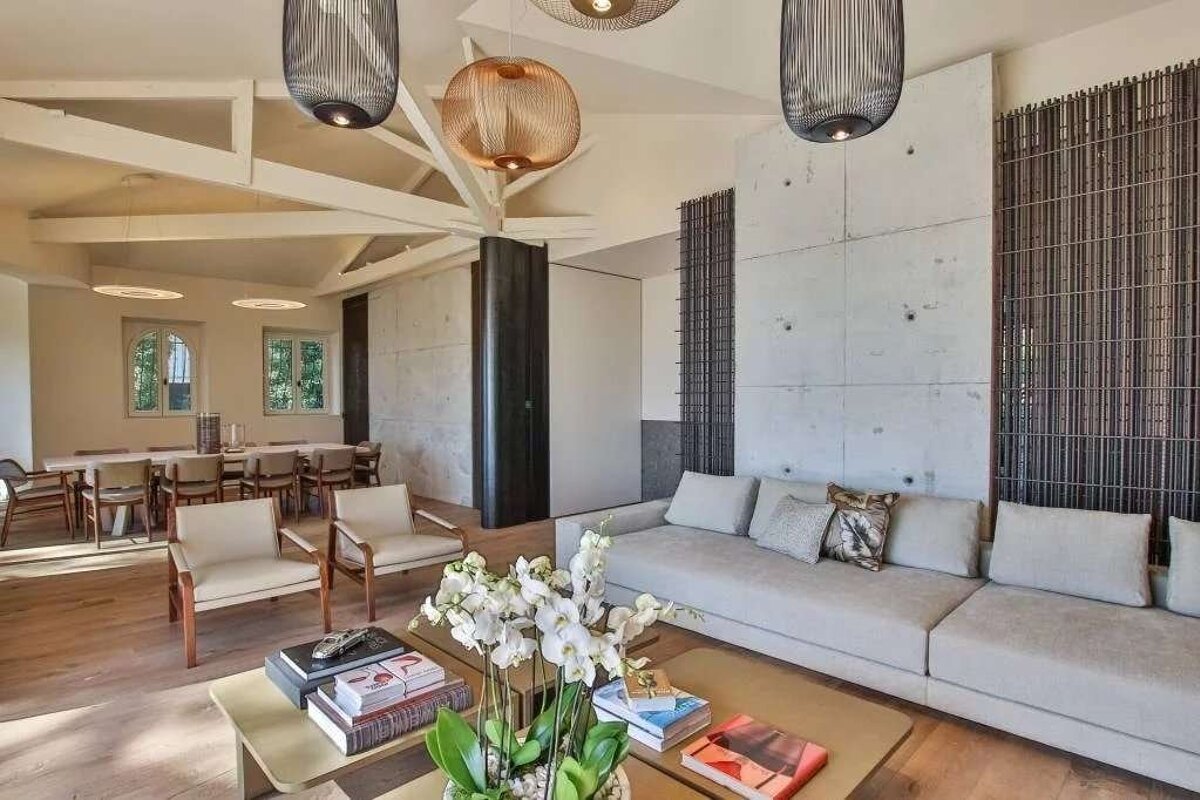 A modern, open-plan living and dining area features concrete walls, exposed beams, sleek furniture, unique hanging lights, and an orchid centerpiece.
