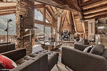 A cozy rustic chalet living room featuring wooden beams, a stone fireplace, plush gray armchairs, and floor-to-ceiling mountain views.