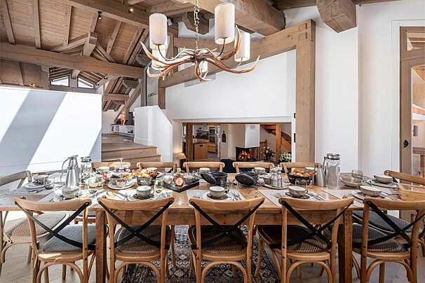 Rustic chalet dining room with a long wooden table set for breakfast, surrounded by cross-back chairs under an antler chandelier.