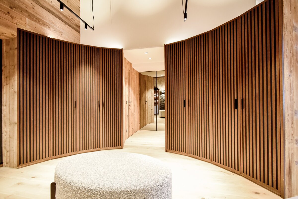 Modern room with curved, vertical-slatted wooden wardrobes, a light wood floor, and a round textured ottoman.