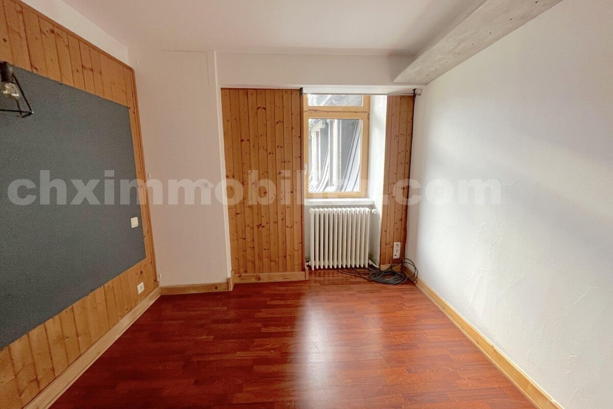 Empty room with dark wood floor, white walls, and a wood-paneled section with a dark board. A window with a radiator is at the back.