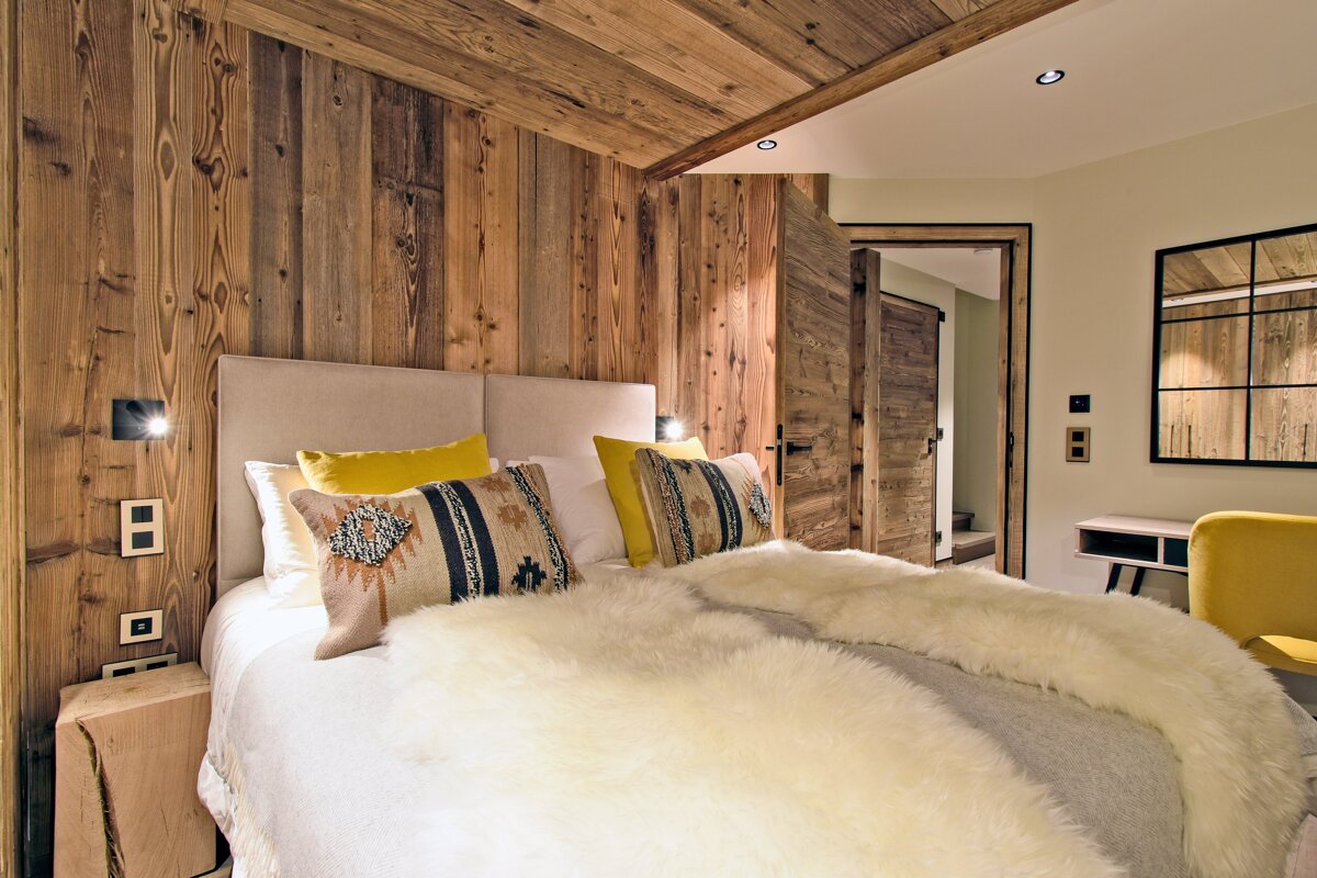 A warm, rustic bedroom with wood-paneled walls/ceiling, a cozy bed featuring a fluffy white throw, and yellow accent pillows/chair.