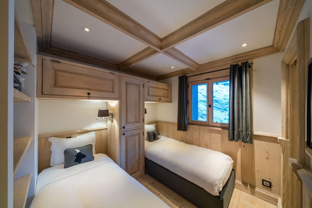A cozy, wood-paneled twin bedroom with built-in storage, exposed ceiling beams, and a window showcasing a snowy outdoor scene.