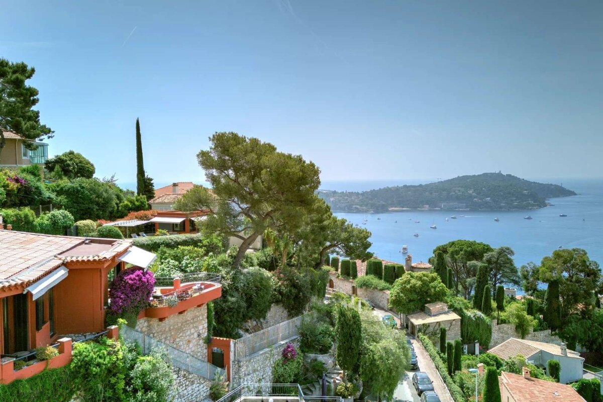 A vibrant coastal landscape features terracotta-roofed houses nestled among lush green hills, overlooking a sparkling blue sea with boats and a distant island.