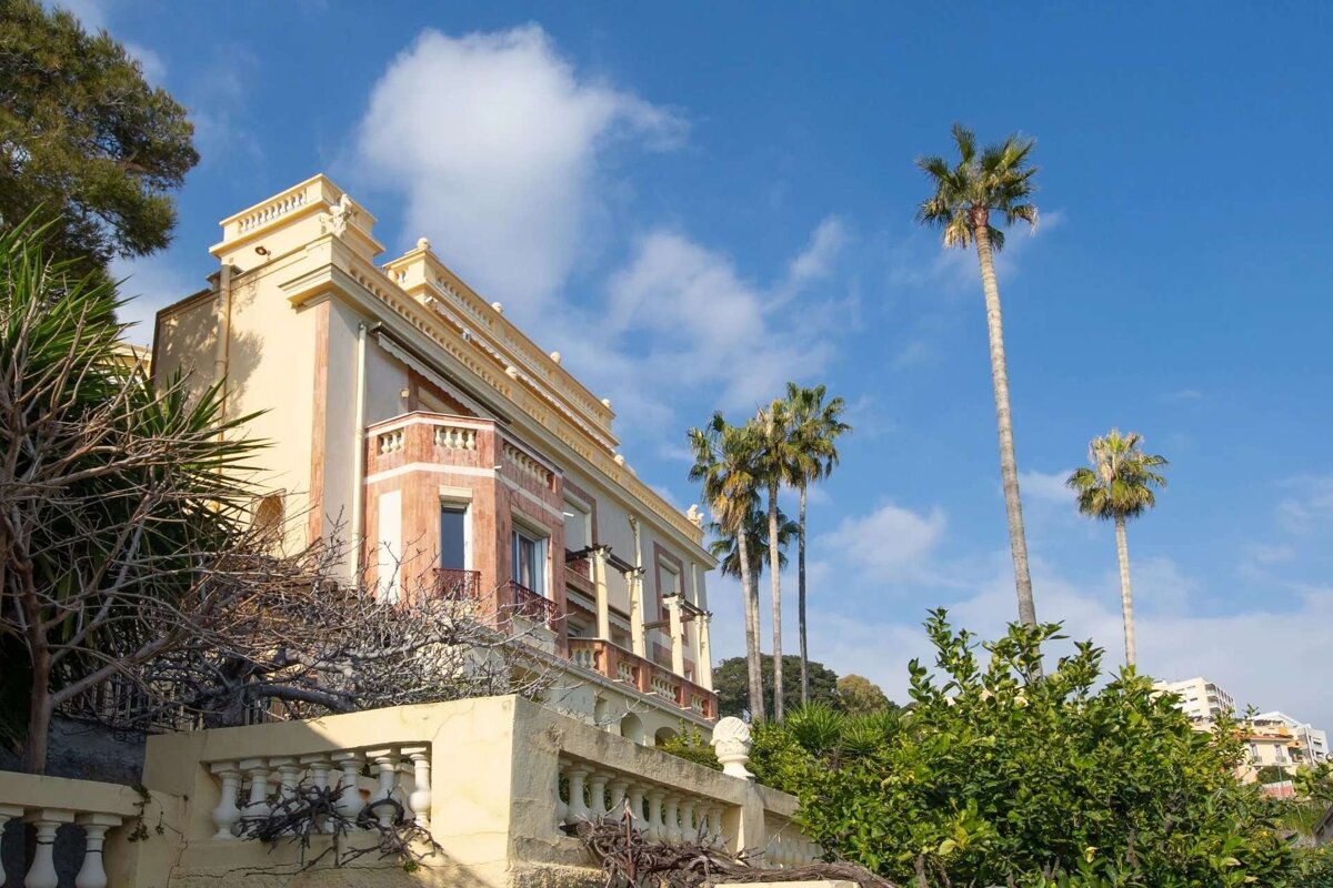 An ornate, multi-story yellow and orange building with intricate details, set against a blue sky with clouds. Tall palm trees and green foliage surround it.