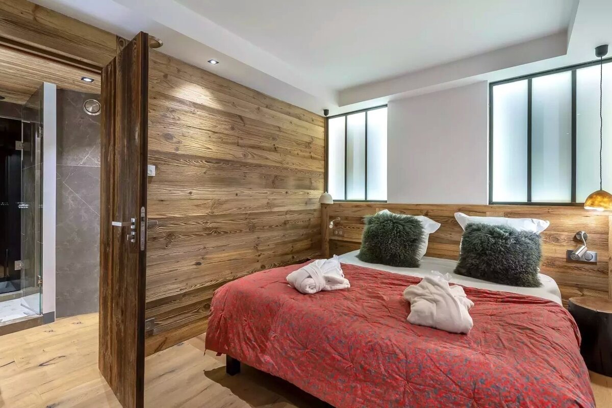 A cozy bedroom features rustic wood paneling, a red bed with fluffy pillows and white bathrobes. A modern shower is visible through a pivoting door.