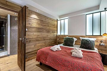 A cozy bedroom features rustic wood paneling, a red bed with fluffy pillows and white bathrobes. A modern shower is visible through a pivoting door.