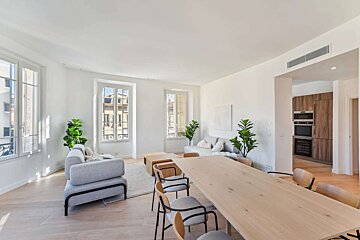 A bright, modern open-plan living/dining area with white walls, light wood floors, grey sofas, large wooden table & chairs, and green plants. Kitchen visible.