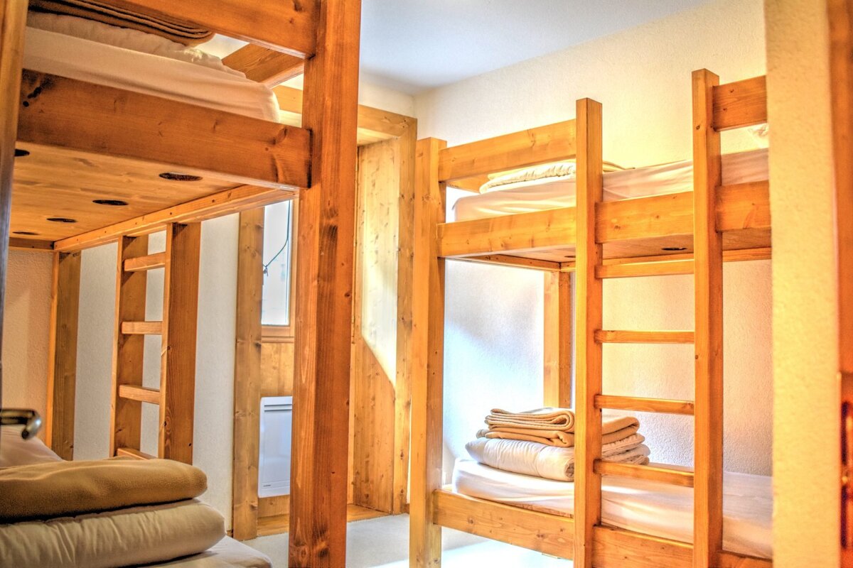 A bright room with several sturdy wooden bunk beds, neatly made with white sheets and folded blankets, suggesting a cozy hostel or dorm.