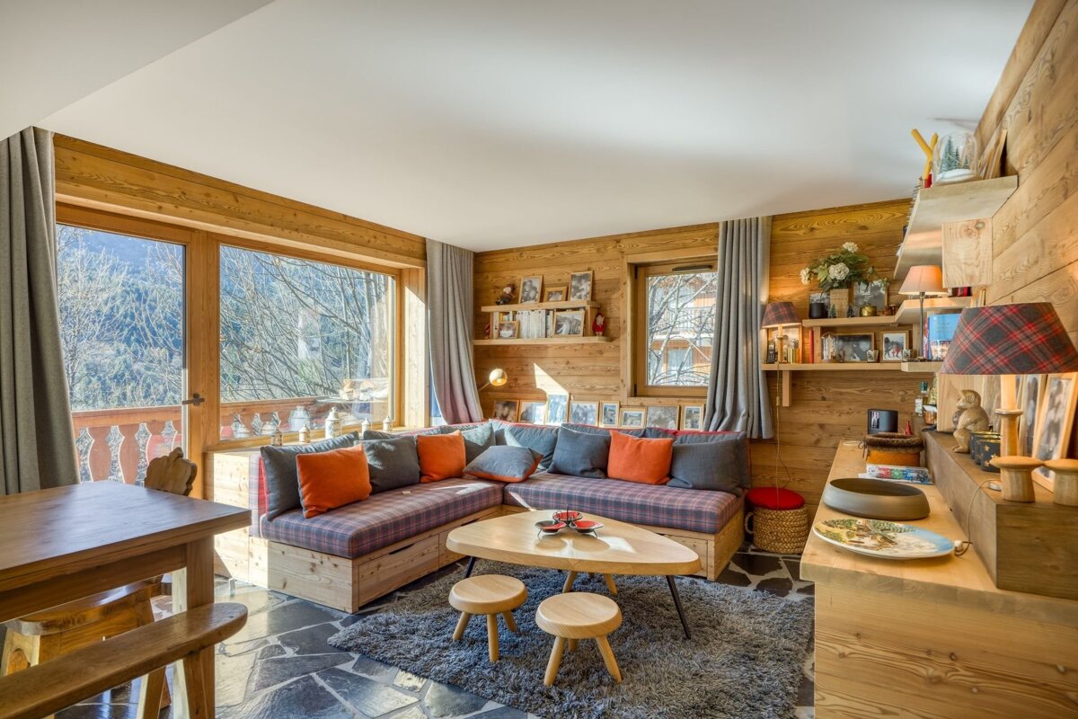 A cozy, wood-paneled chalet living room featuring a plaid sectional, wooden furniture, and large windows with a view of winter trees, creating a warm ambiance.