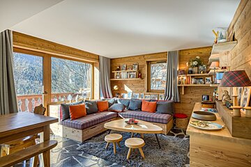 A cozy, wood-paneled chalet living room featuring a plaid sectional, wooden furniture, and large windows with a view of winter trees, creating a warm ambiance.