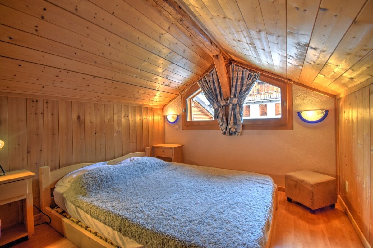 A cozy, wood-paneled attic bedroom features a bed with a fluffy grey blanket, a window with checkered curtains, nightstands, and warm wall lights.