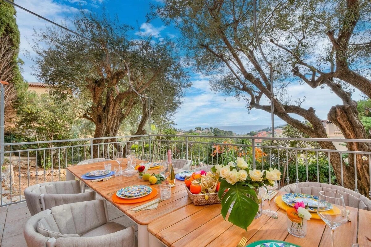 An outdoor wooden dining table set for a meal on a balcony overlooking trees and a distant sea view under a blue sky.