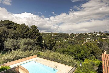 An aerial view shows a swimming pool overlooking a green, tree-covered hillside dotted with houses, under a cloudy blue sky.
