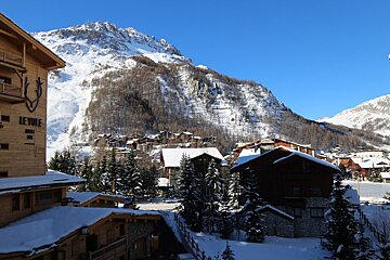 Snow-covered wooden chalets in a mountain ski resort with pine trees and a towering snow-capped peak under a clear blue sky.