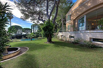 Luxurious house, lush garden with large trees, fountain, and distant swimming pool under a blue sky.