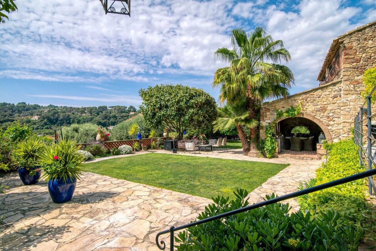 Mediterranean stone villa courtyard with a lush lawn, palm tree, paved patio, and scenic landscape under a bright, cloudy sky.