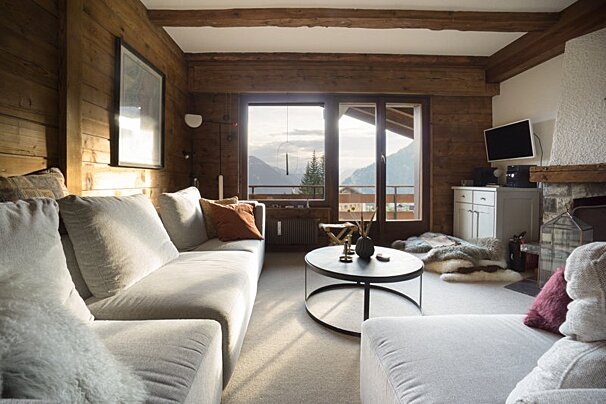 Cozy alpine chalet living room with wooden walls, light gray sofas, a round coffee table, and large windows overlooking mountains.