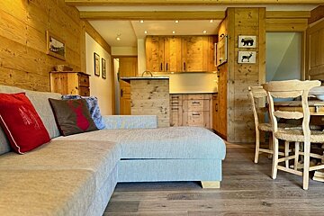 Cozy cabin interior with a grey sectional sofa, wood-paneled walls, kitchen cabinets in the background, and a rustic dining area.