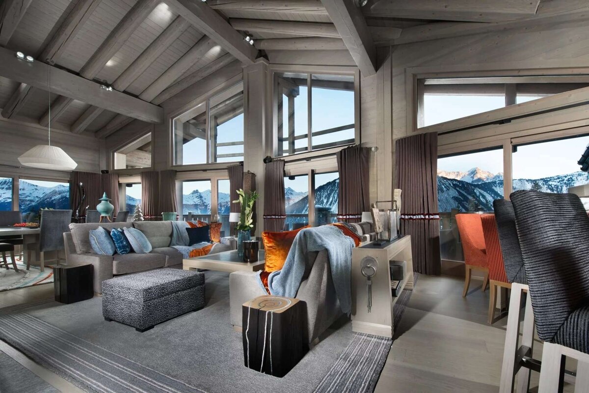Modern luxury ski chalet interior featuring high wood-beamed ceilings, large windows with panoramic snowy mountain views, and comfortable, chic furnishings.