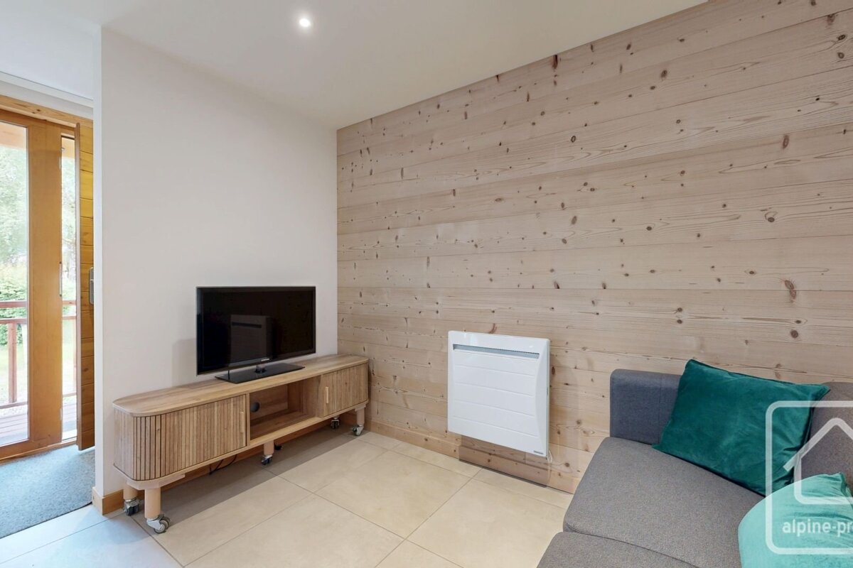 A modern room featuring a light wood-paneled wall, a grey sofa with a teal pillow, and a TV on a wooden console. Light tiled floor and a doorway on the left.