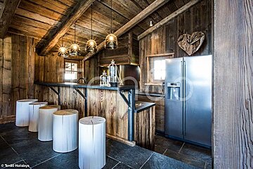 Rustic wooden cabin kitchen featuring a bar with white log stools, hanging pendant lights, and a stainless steel refrigerator.
