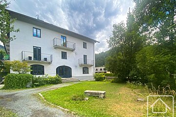 A white multi-story building with balconies overlooks a grassy yard. Trees, mountains, and a cloudy sky complete the outdoor scene.