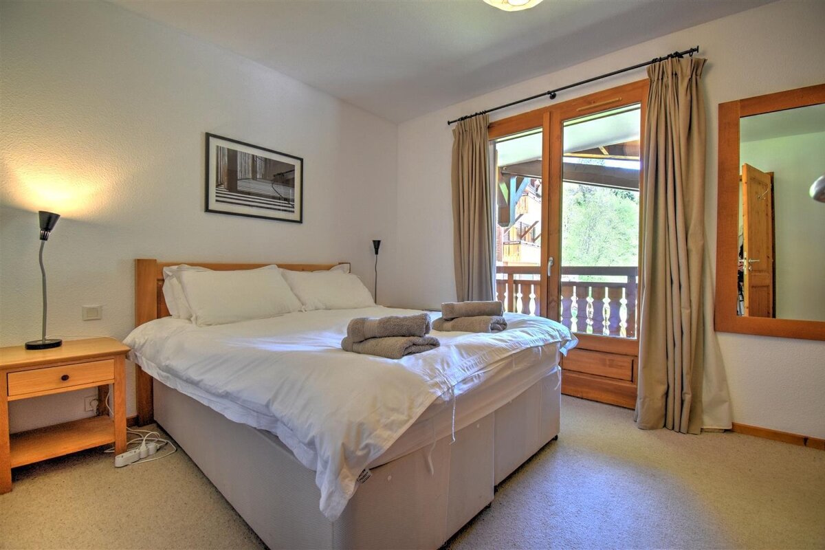 A cozy bedroom features a large bed with white linens, wooden headboard and nightstand, and a balcony door with curtains. A mirror hangs on the wall.