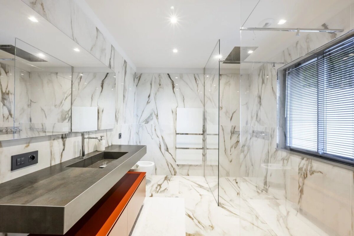 Elegant modern bathroom with white marble surfaces, glass shower, sleek dark sink vanity with an orange base, and a window with blinds.