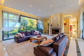 Luxurious living room with brown leather sofas, a grand fireplace, and large windows revealing a lush outdoor garden. Features yellow walls and elegant lighting.