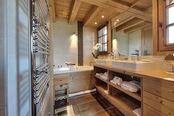 A warm, rustic chalet-style bathroom features wooden ceilings and vanity, dual sinks, a large mirror, a bathtub, and a chrome towel warmer.