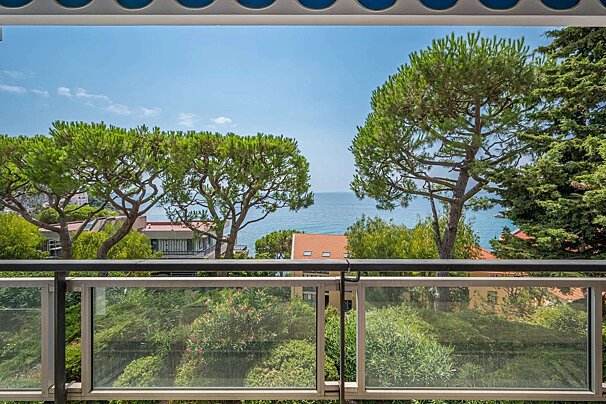 A balcony overlooks a calm blue sea through lush green pine trees and foliage. Houses with red roofs are visible below, all under a clear sky.