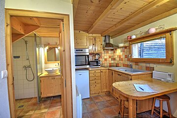 A cozy, rustic studio apartment with a wooden kitchen featuring appliances and a dining table, connected to a tiled bathroom with a shower and sink.