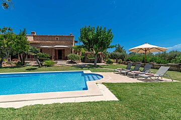 A charming stone house with a private swimming pool, green grass, sun loungers, and a beige umbrella under a bright blue sky.
