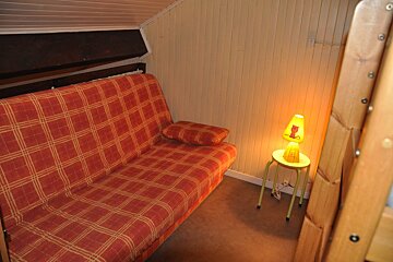A cozy room features a red plaid futon, light paneled walls, and a glowing yellow lamp with a cat design on a stool. Part of a wooden bunk bed is visible.