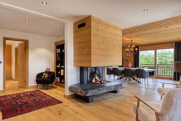 A modern open-plan chalet with a central wooden fireplace, light wood floors, and a dining area overlooking a mountain view. Cozy and rustic elegance.