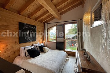 A cozy wood-paneled bedroom features an angled ceiling, a large bed with modern pillows, a portrait, and a window opening to a green view.